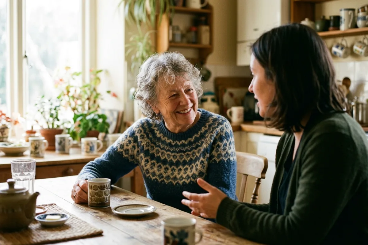 An older adult laughing during a relaxed conversation at a sunlit kitchen table with family nearby